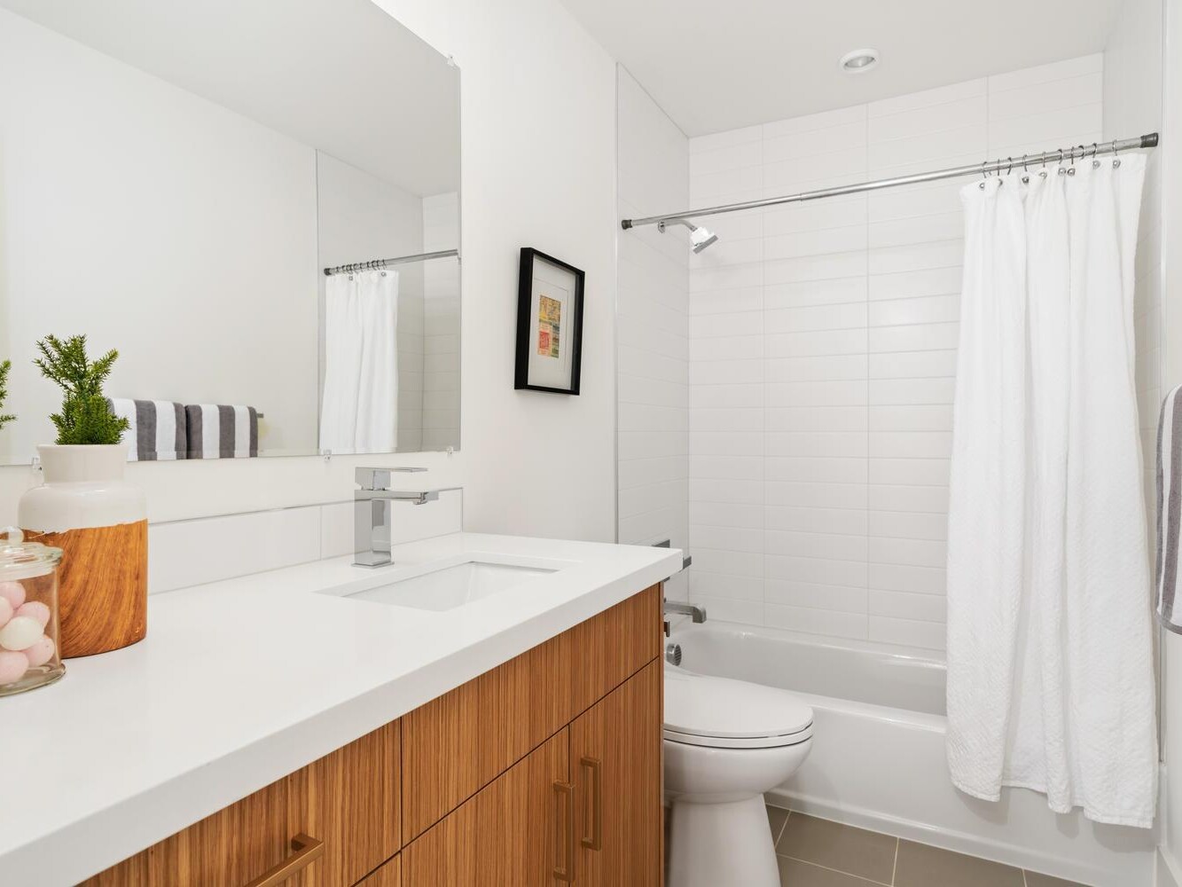 Modern bathroom with a white countertop, wood vanity, sink, toilet, and a bathtub with a white shower curtain. Towels, a framed picture, and small plants decorate the space for a clean, minimalist look.