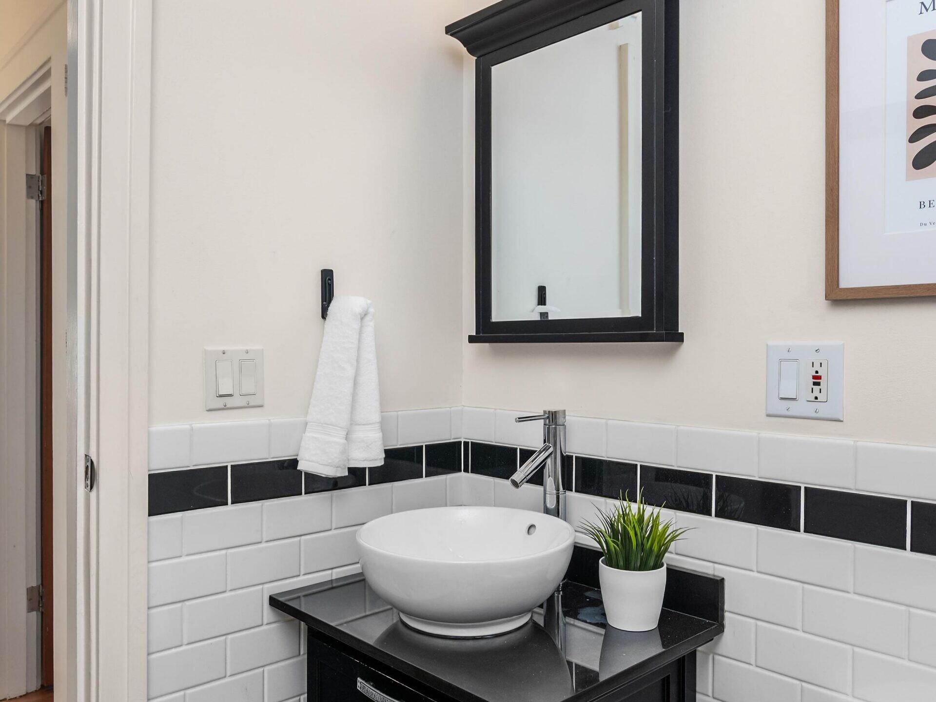 Modern bathroom with a black vanity, white vessel sink, wall-mounted mirror cabinet, towel hanging on a hook, potted plant, and tiled white walls with a black accent stripe.