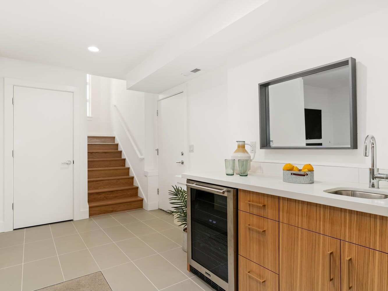Modern kitchenette with wooden cabinets, a small sink, a wine fridge, a wall mirror, and decorative vases. Stairs with wooden steps lead up, and white doors are visible in the bright, minimalist space.