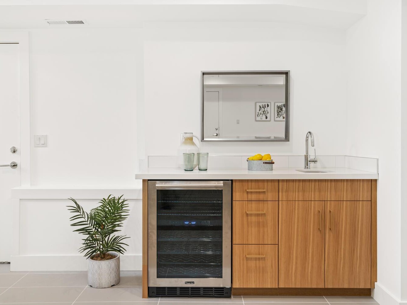 A modern kitchenette with wood cabinets, a small sink, a countertop holding a bowl of lemons and glasses, a built-in mini fridge, a potted plant on the floor, and a wall mirror above the sink.