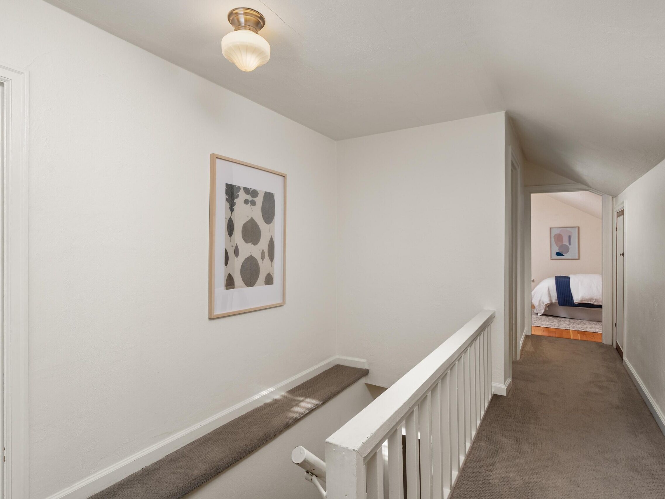 A hallway with beige carpet, white walls, framed abstract art, a white railing, and a ceiling light leads to a bedroom with a made bed visible in the background.