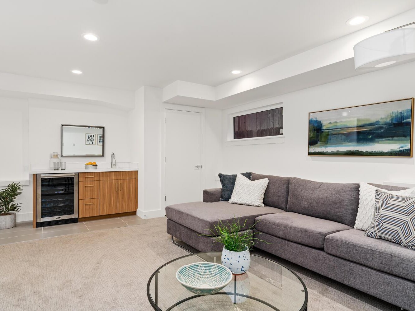 Modern living room with a gray sectional sofa, glass coffee table with a potted plant, wall art, and a small kitchenette featuring wood cabinets, a sink, and a mini-fridge. The room is well-lit and decorated in neutral tones.