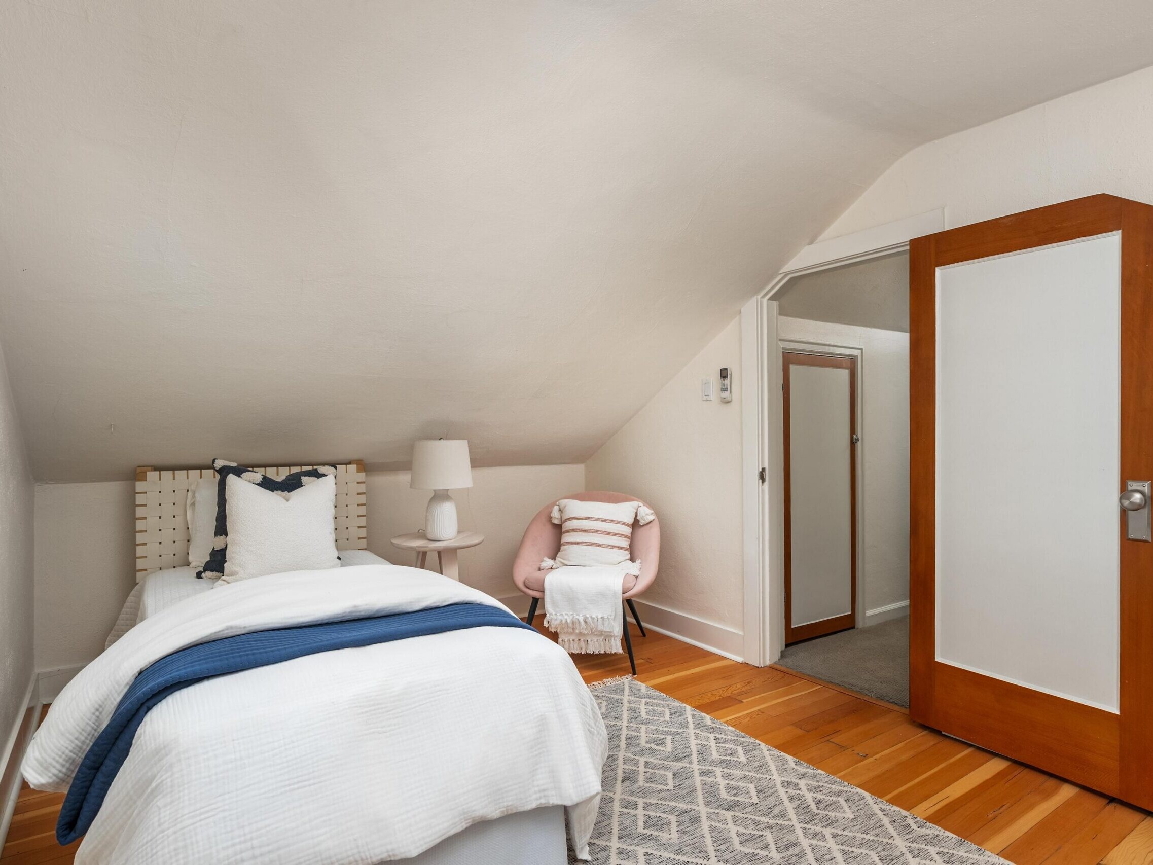 A small attic bedroom with sloped ceiling, featuring a single bed with white bedding, a light pink chair with a striped pillow, a white lamp on a nightstand, wooden floor, and an open wooden door.