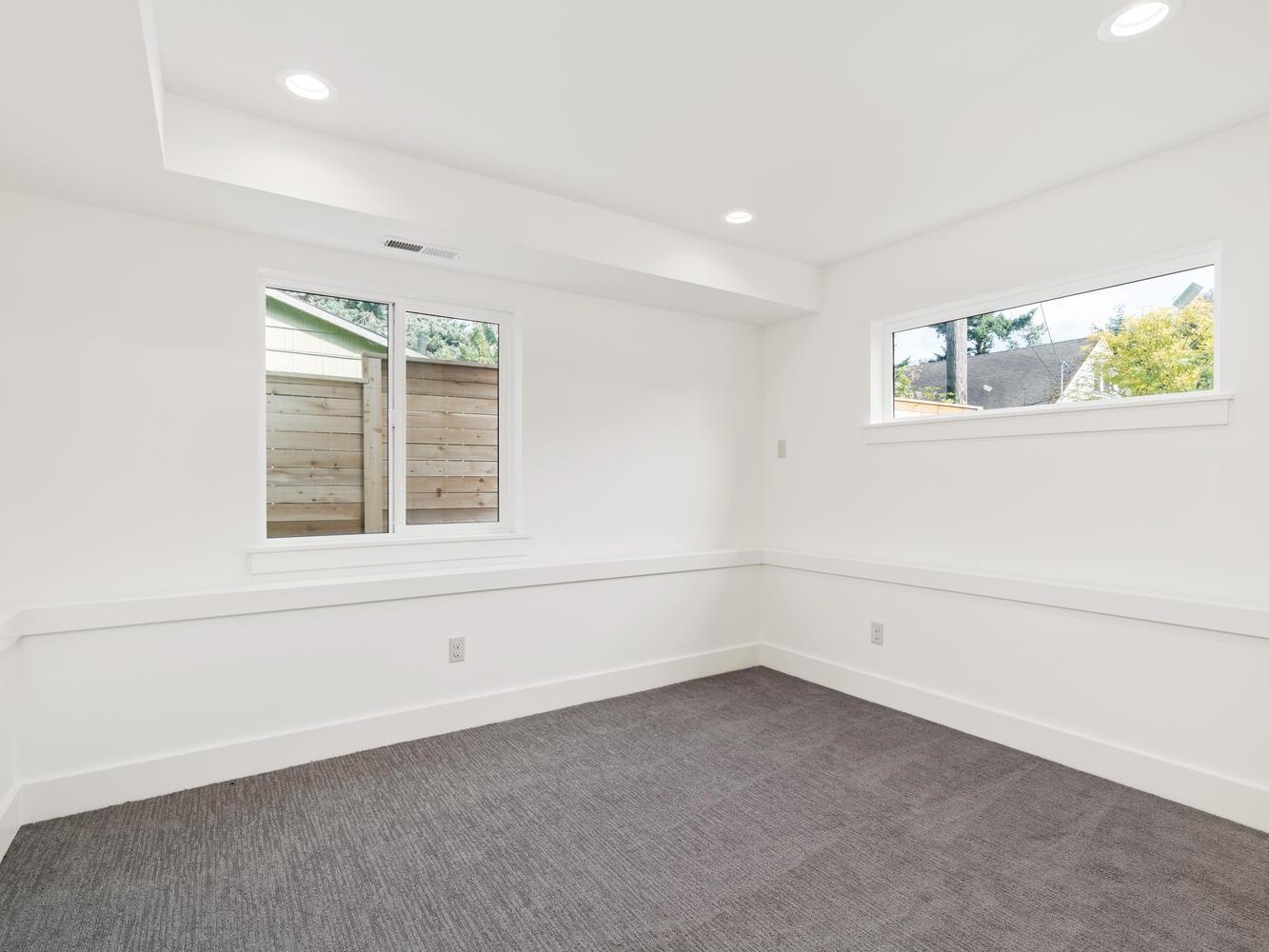 A bright, empty room with white walls, two rectangular windows, gray carpet flooring, and recessed ceiling lights. There is a wooden fence and greenery visible outside the windows.