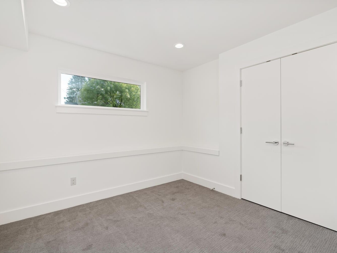 A small, empty room with white walls, gray carpet, a narrow horizontal window showing greenery outside, and double white doors. The space is clean and well-lit with recessed ceiling lights.