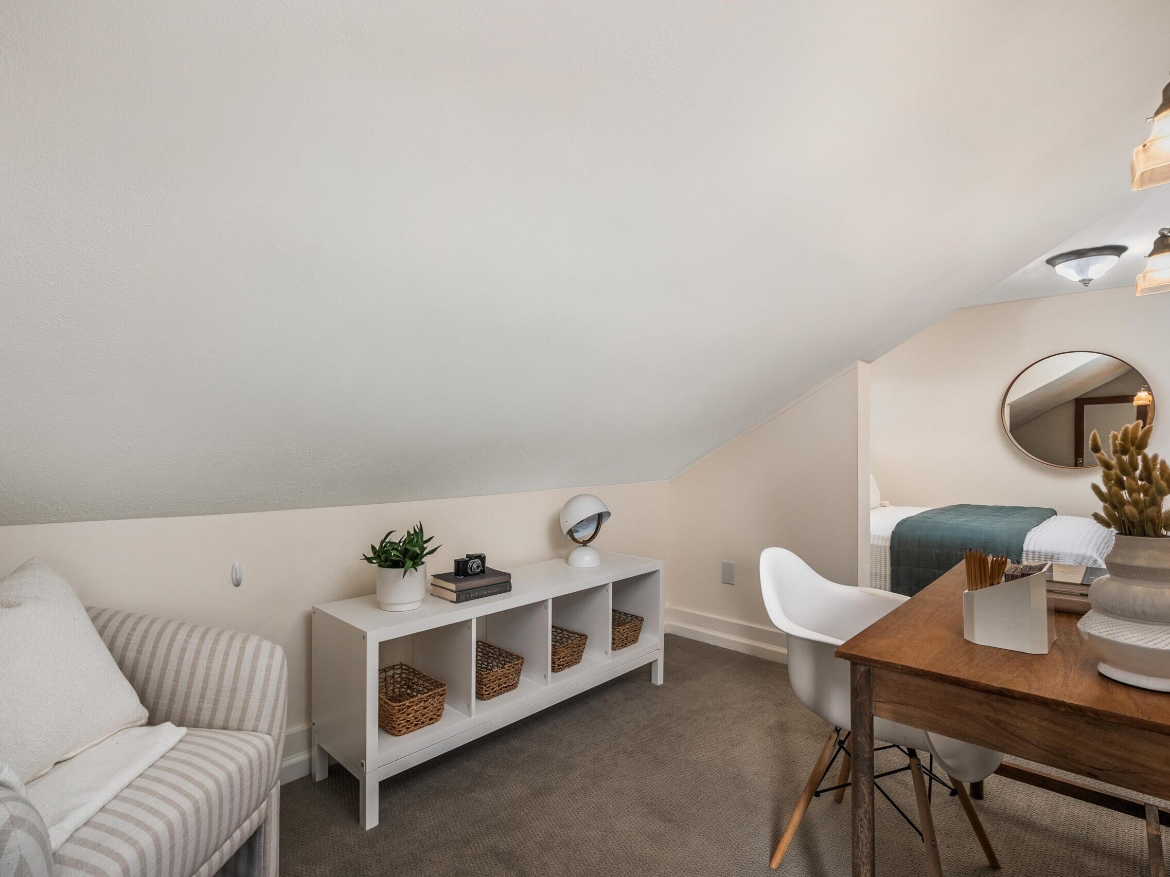 A cozy attic room with a sloped ceiling, featuring a striped armchair, a white shelf with baskets, a desk with a chair, and a bed in the background with a round mirror above it.