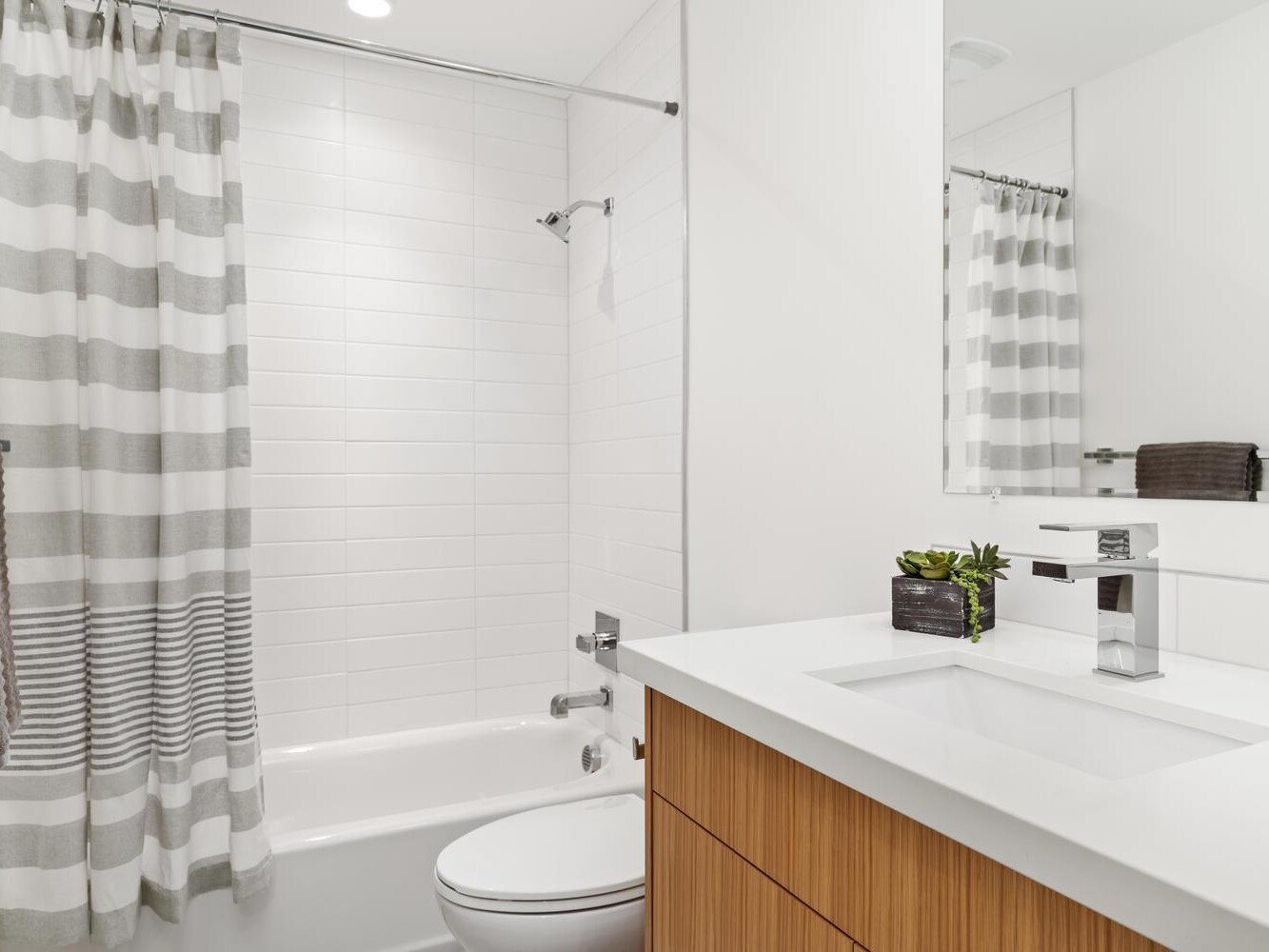 Modern bathroom with white walls, a wooden vanity, a rectangular sink, a large mirror, and a bathtub with a striped gray and white shower curtain. A small plant and rolled towel sit on the counter.