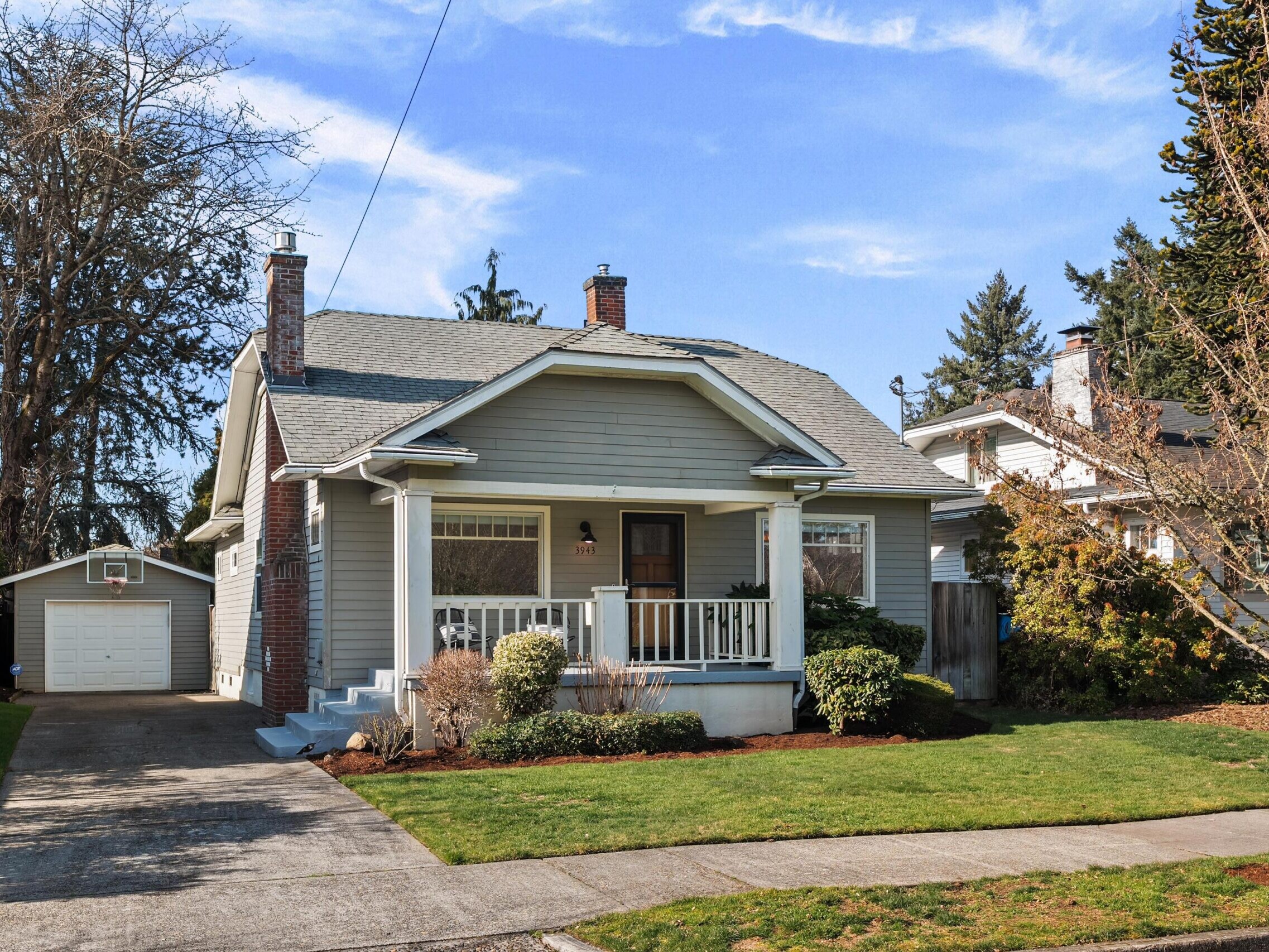 A gray single-story house with a covered front porch, white trim, and a small lawn. There is a driveway leading to a detached garage and leafless trees surrounding the property on a sunny day.