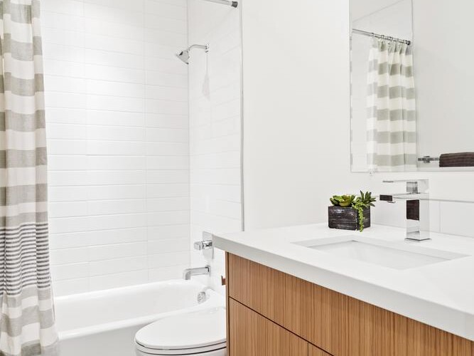 Modern bathroom with a wood vanity, white countertop, rectangular mirror, toilet, and a bathtub with a white and gray striped shower curtain. Walls are white with light gray tile floor. Small plant on the counter.