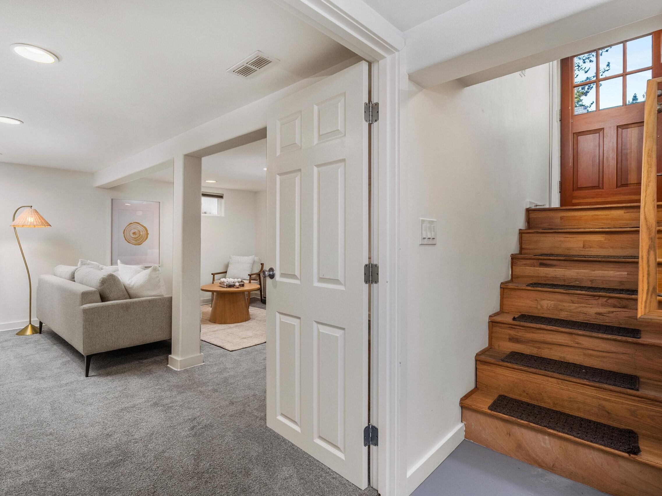 A carpeted basement with a gray sofa, a floor lamp, a small table, and a white door, next to a wooden staircase leading up to a door with glass panes. The walls are white and the space is well-lit.