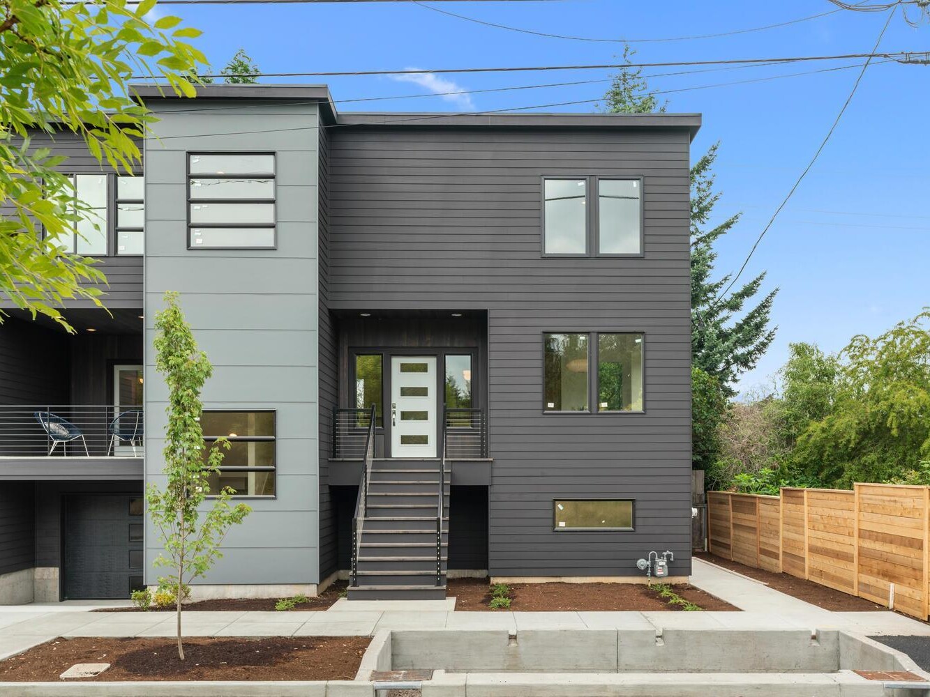 Modern two-story gray house with large windows, a front door reached by stairs, a small balcony on the left, young trees in front, and a wooden privacy fence on the right, under a clear blue sky.