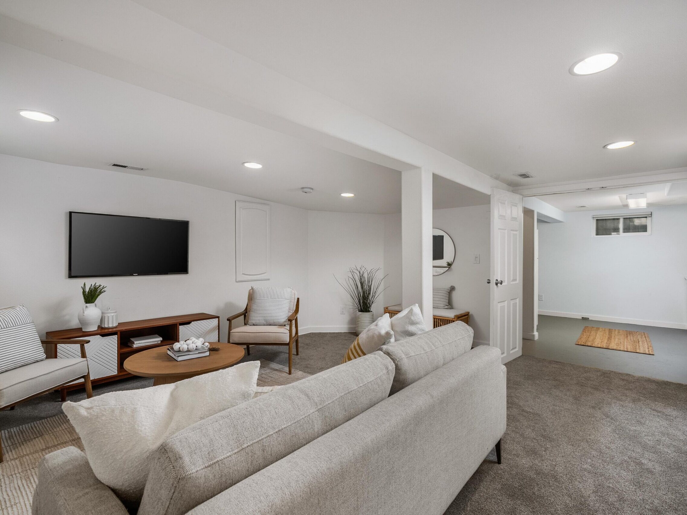 A modern basement living area with light gray carpet, a beige sofa, a round wooden coffee table, two armchairs, a wall-mounted TV, and neutral decor, featuring white walls and recessed ceiling lights.