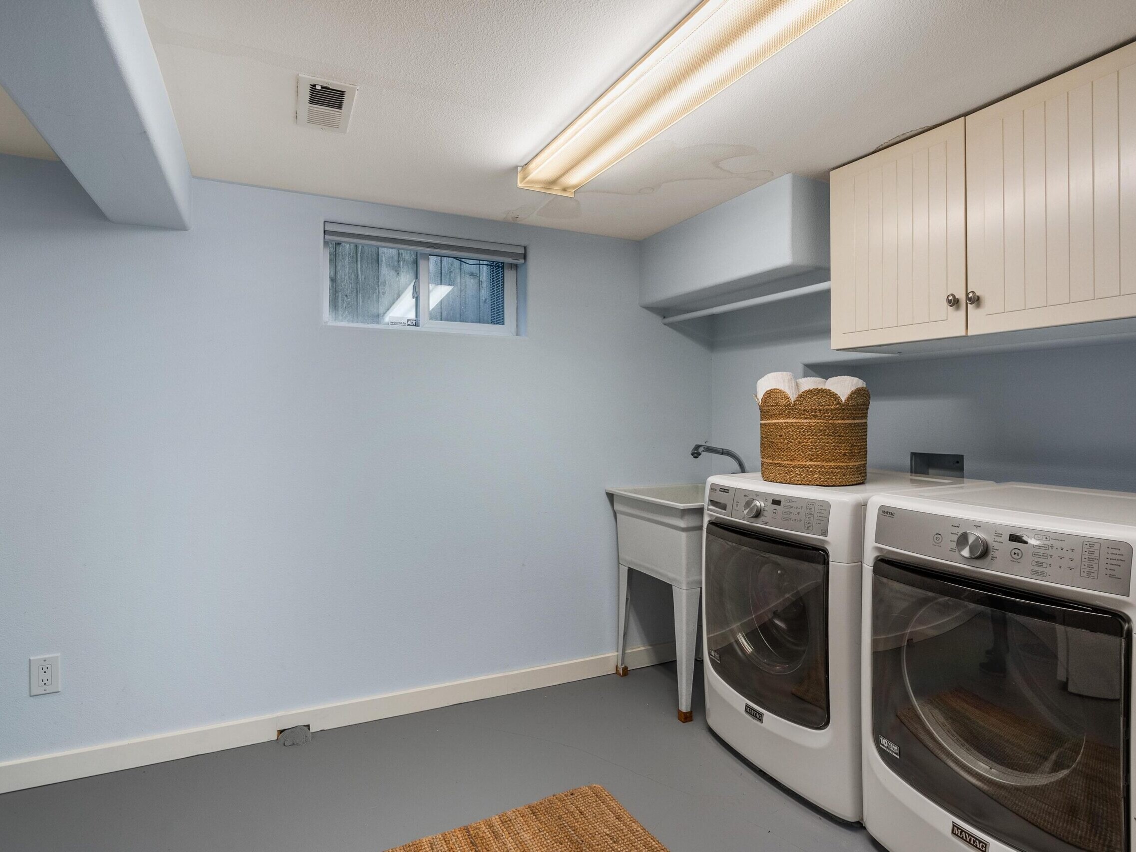 A clean laundry room with light blue walls, a washer and dryer side by side, a laundry basket with towels on top, a utility sink, overhead cabinets, a window, and a fluorescent ceiling light.