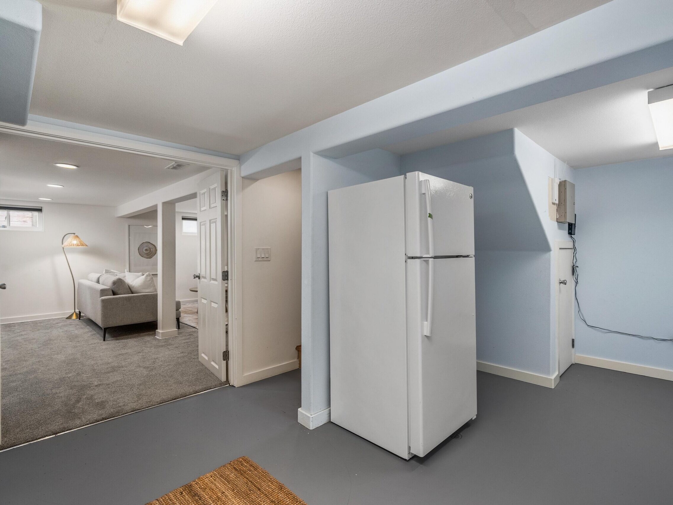 A basement room with light blue walls and gray flooring features a white refrigerator. An open doorway leads to a carpeted living area with a sofa and floor lamp. Ceiling lights brighten both rooms.