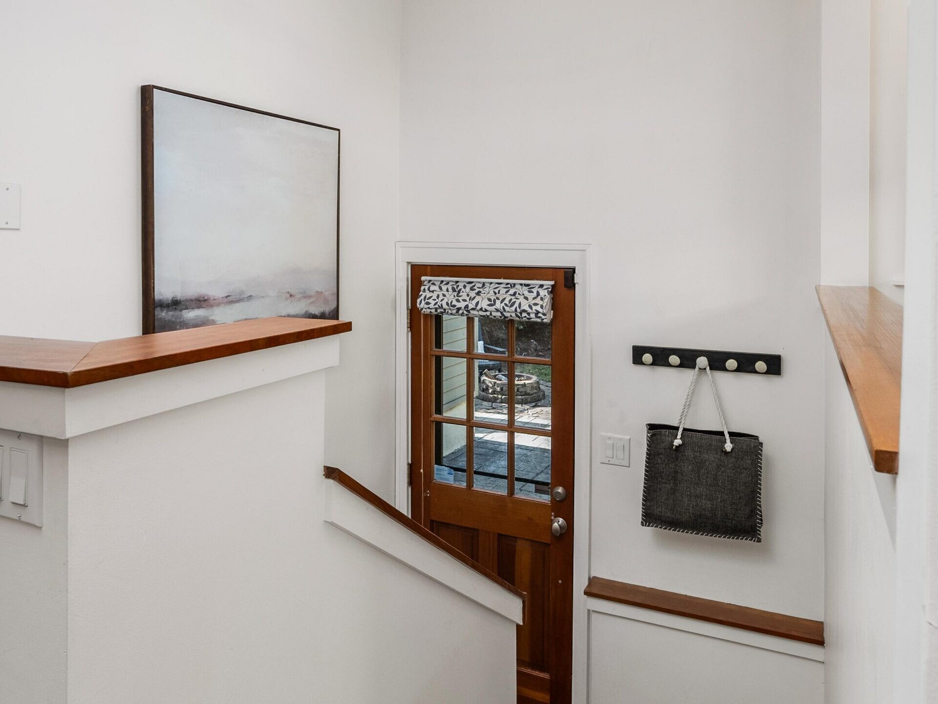 A small, tidy entryway with wooden stairs leading down to a glass-paneled wooden door. A modern painting hangs on the wall, and a coat rack with a bag and basket is mounted by the door. The space is well-lit and minimalistic.
