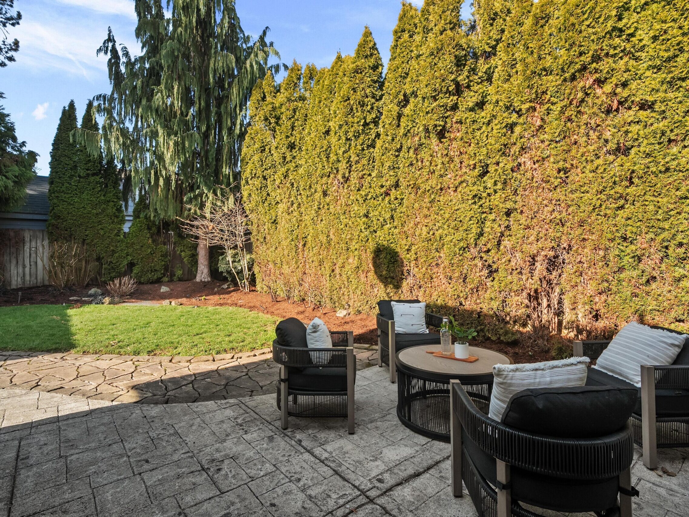 A backyard patio with black cushioned chairs and a round table overlooks a small lawn bordered by tall evergreen trees and a wooden fence under a clear sky.