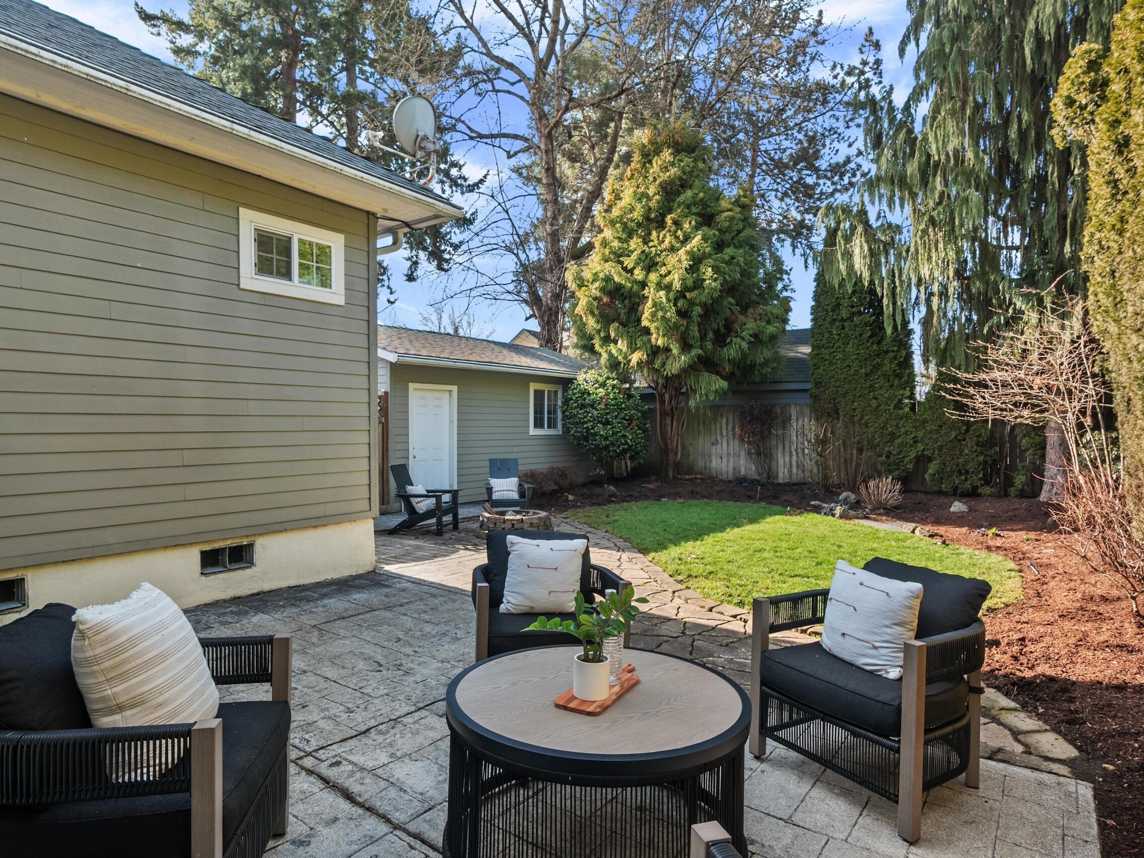 A cozy backyard patio with three black cushioned chairs around a round table, potted plant centerpiece, concrete paving, green grass, and tall trees and shrubs along the fence and house.