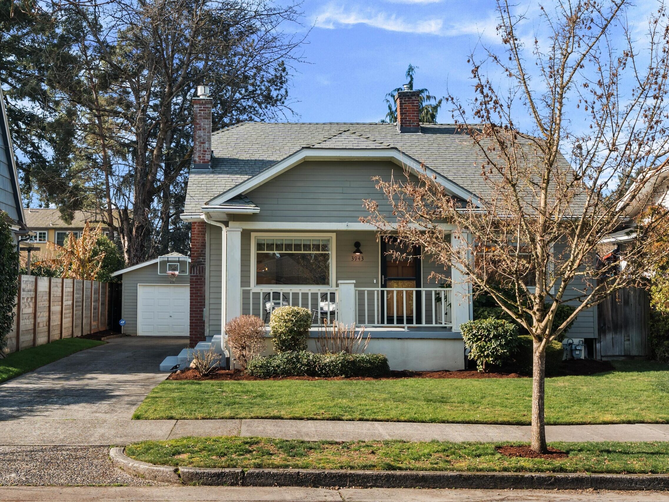 A small, light blue house with a covered front porch, white trim, and a neatly landscaped yard. A tree without leaves stands in the front lawn. There is a driveway on the left leading to a detached garage.