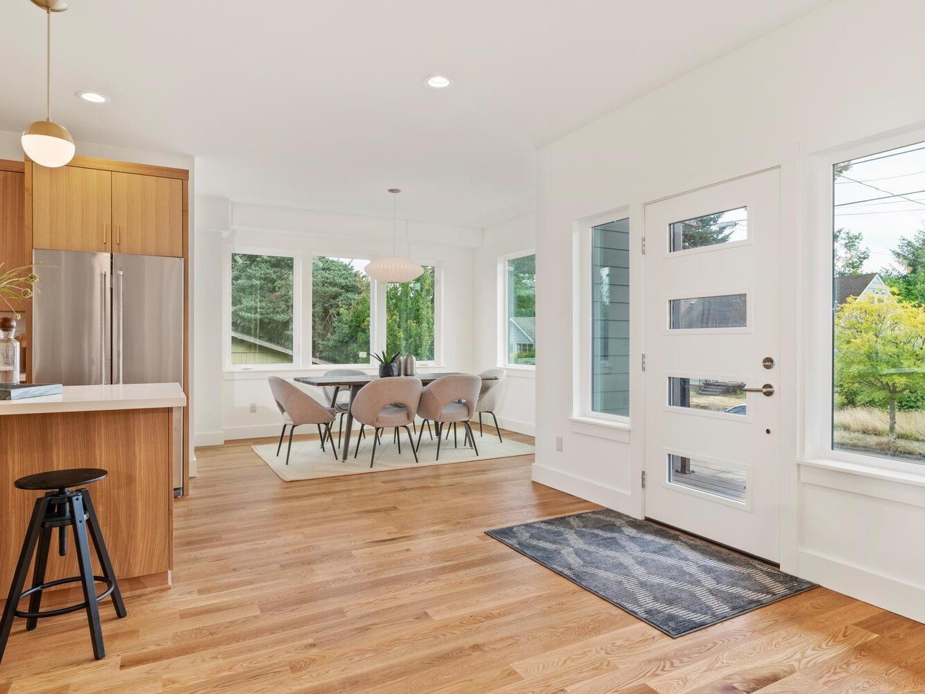 Bright, modern kitchen and dining area with wood floors, light wood cabinets, a kitchen island with a black stool, large windows, and a dining table with gray chairs near the back, letting in natural light.
