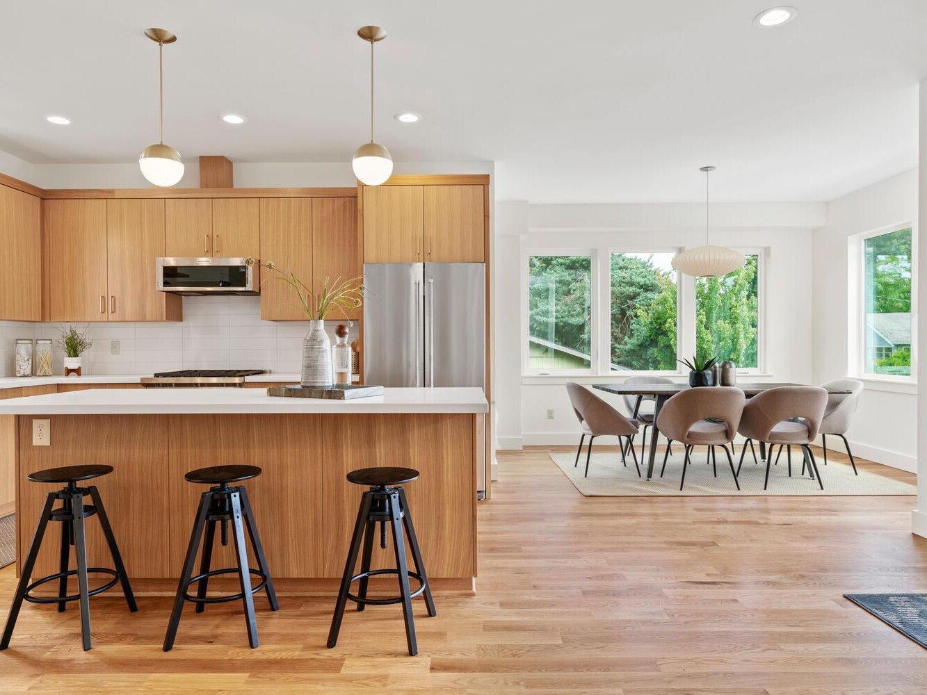 Modern kitchen with light wood cabinets, white countertops, three black barstools, stainless steel appliances, and a dining area with a round table and six gray chairs by large windows with green outdoor views.