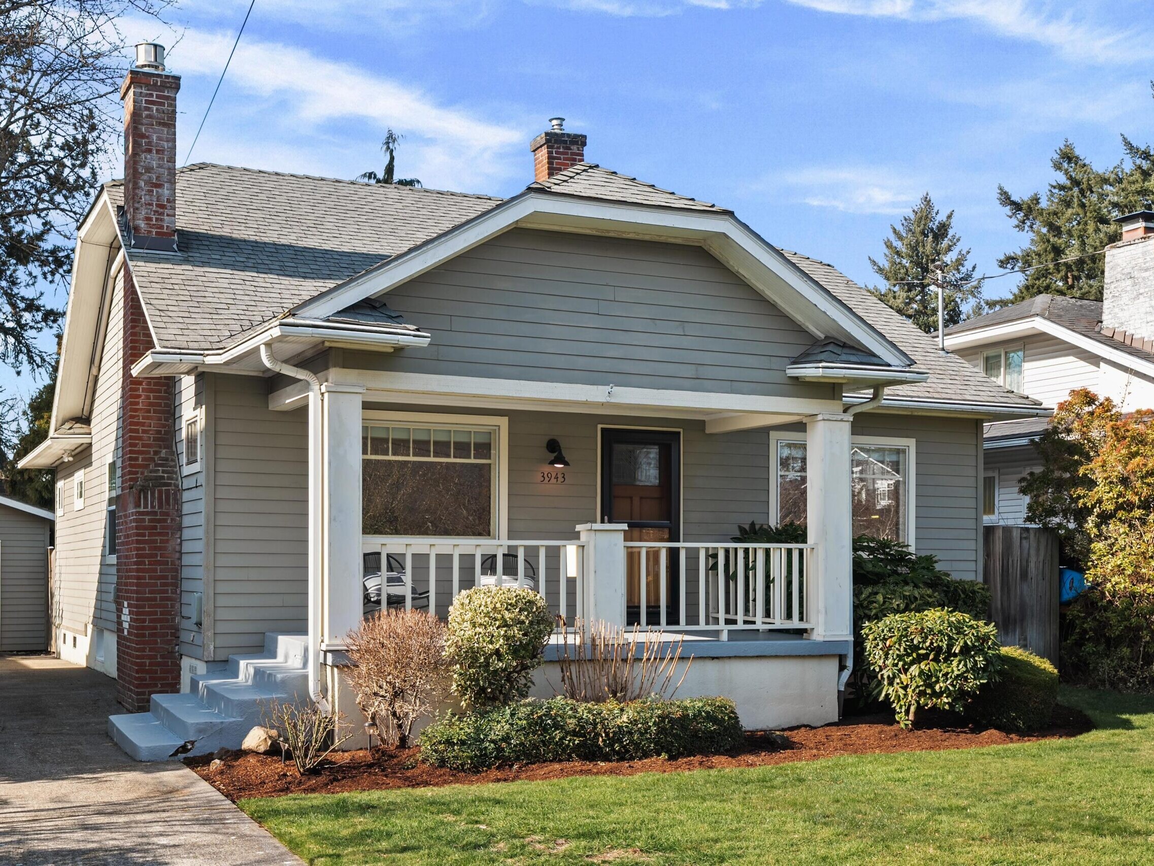 A single-story gray house with a front porch, white trim, and brick chimney, surrounded by green grass and shrubs. A driveway leads to a detached garage, and trees are visible in the background under a blue sky.