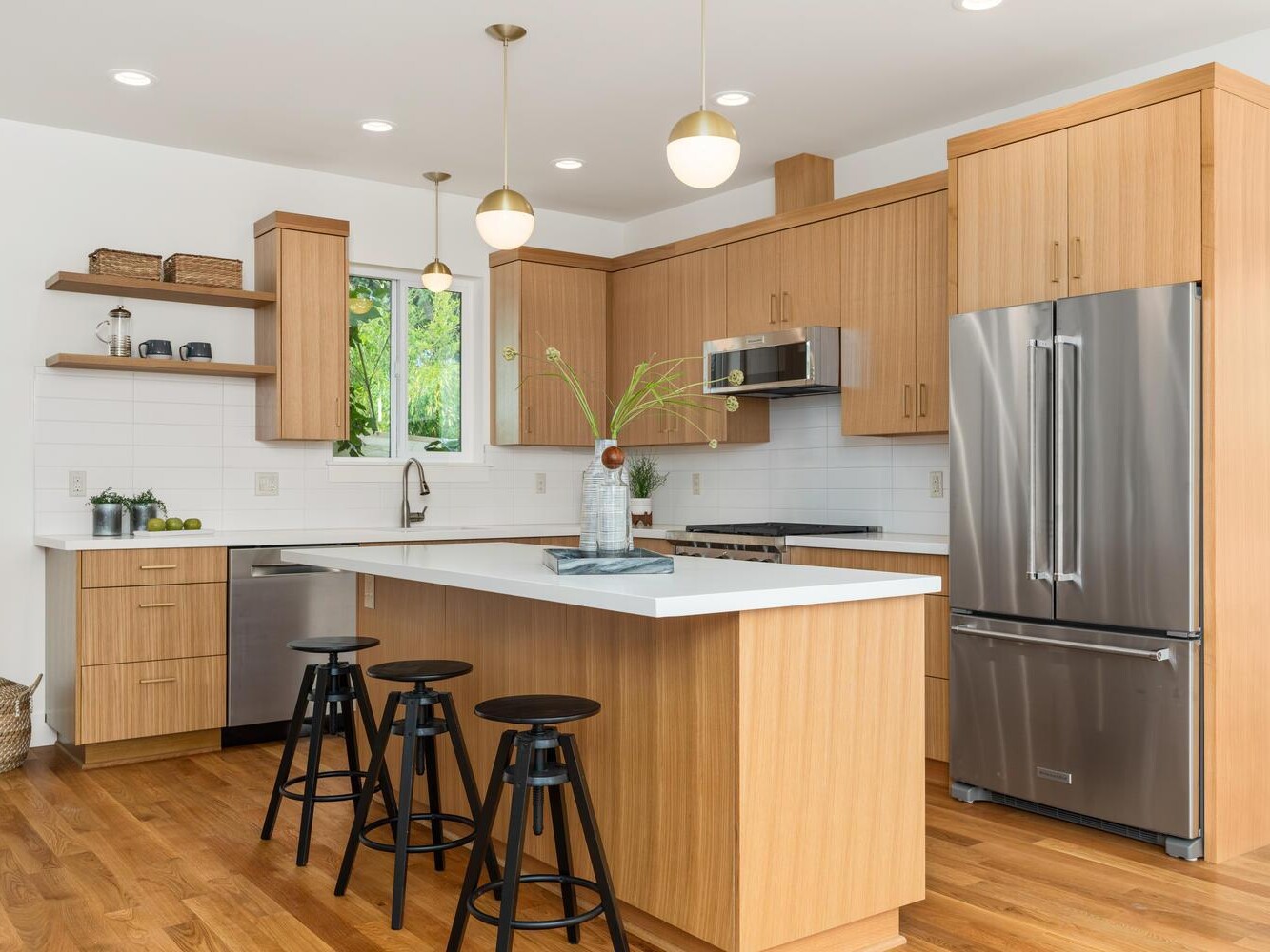 Modern kitchen with light wood cabinets, stainless steel appliances, a center island with three black stools, white countertops, open shelves, a vase with greenery, and hardwood floors.