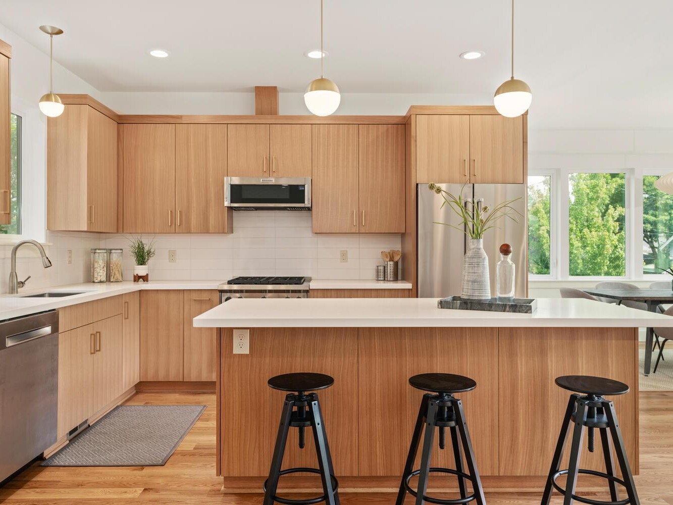 Modern kitchen with light wood cabinets, white countertops, stainless steel appliances, and three black bar stools at an island. Pendant lights hang above, and large windows bring in natural light.