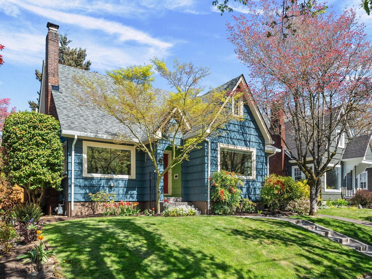 A charming blue house with white trim, a pitched roof, and a green front door. The yard features a well-kept lawn, blooming flowers, shrubs, and trees with spring foliage. Other houses are visible on either side.