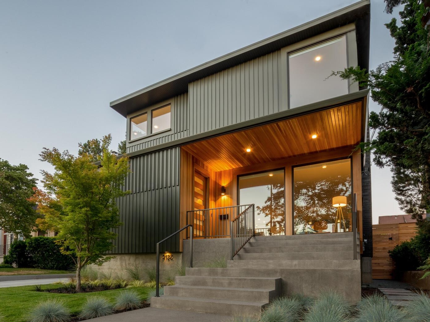 Modern two-story house with large windows, wood accents under the porch roof, concrete steps leading to a glass front door, landscaped yard with ornamental grass, and trees surrounding the property at dusk.