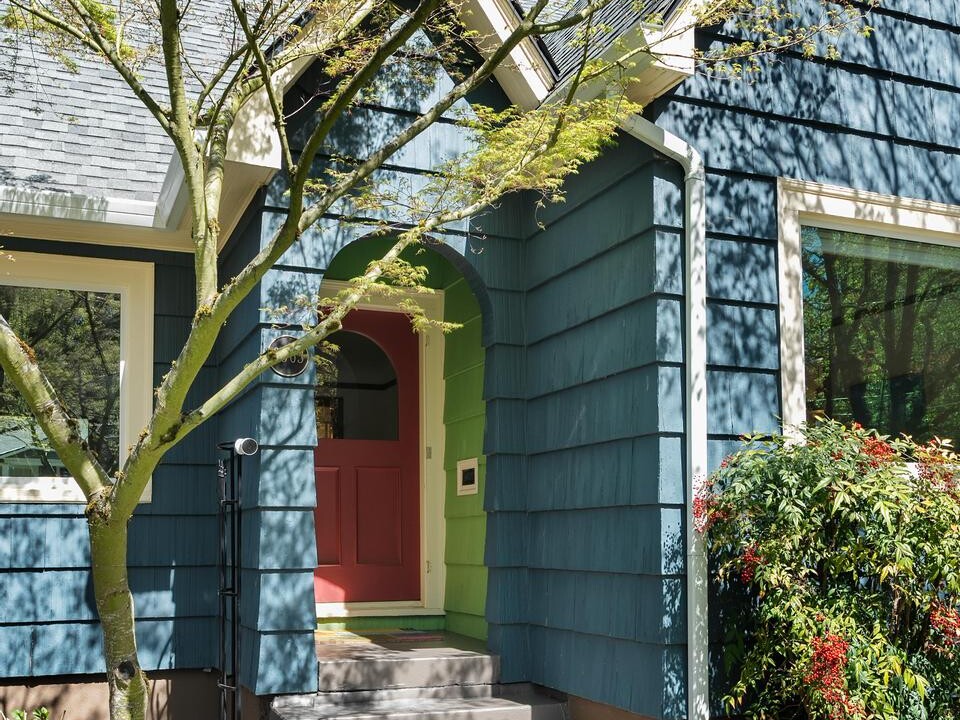 A blue house with shingle siding, a red front door, and arched entryway. Steps lead up to the door, with flowering plants and a small tree in the landscaped front yard. Sunlight casts shadows on the house.