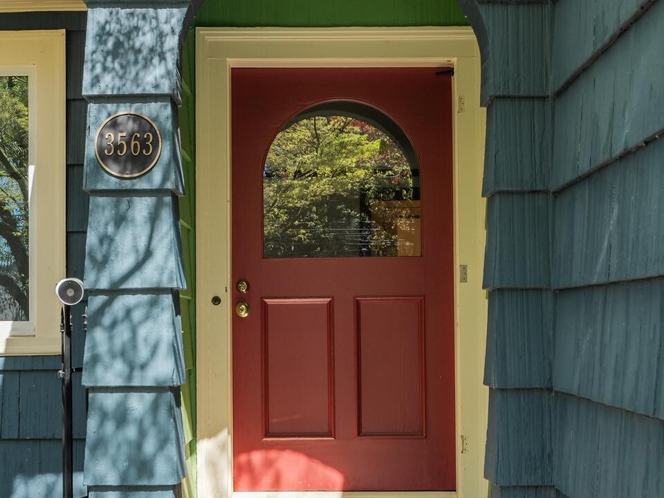 A red front door with a glass window is set in a blue-paneled house under a green arched entryway. The house number 3563 is mounted to the left, and colorful welcome mats are on the steps.