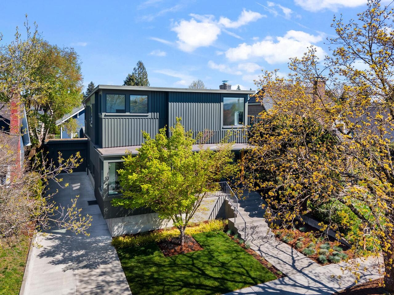 A modern two-story house with dark siding, large windows, and a flat roof stands amid green trees and landscaped lawns on a sunny day, with broad driveways and walkways leading up to the entrance.