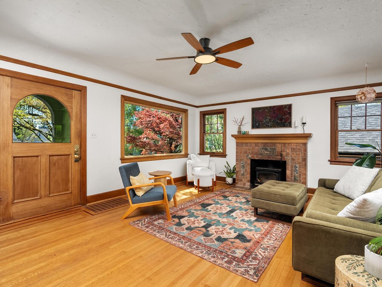 A bright living room with wood floors, a green sofa, patterned rug, armchairs, brick fireplace, large windows showing trees, and a wooden front door. A ceiling fan is centered on the white ceiling.