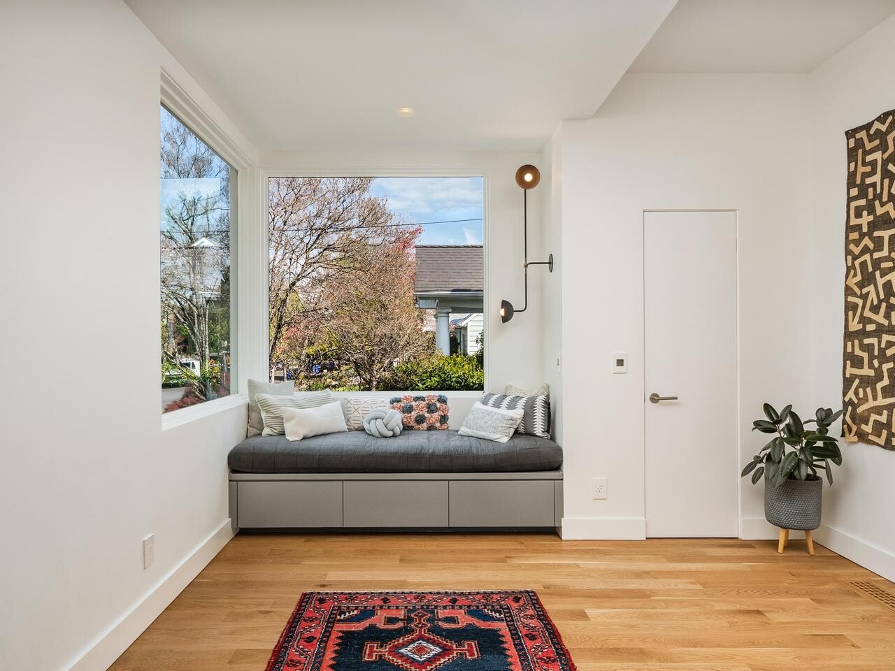 A cozy modern nook with a gray cushioned window seat, patterned pillows, and a wall sconce. Large windows let in natural light. A potted plant, wall art, and a red patterned rug add warmth to the clean, minimalist space.