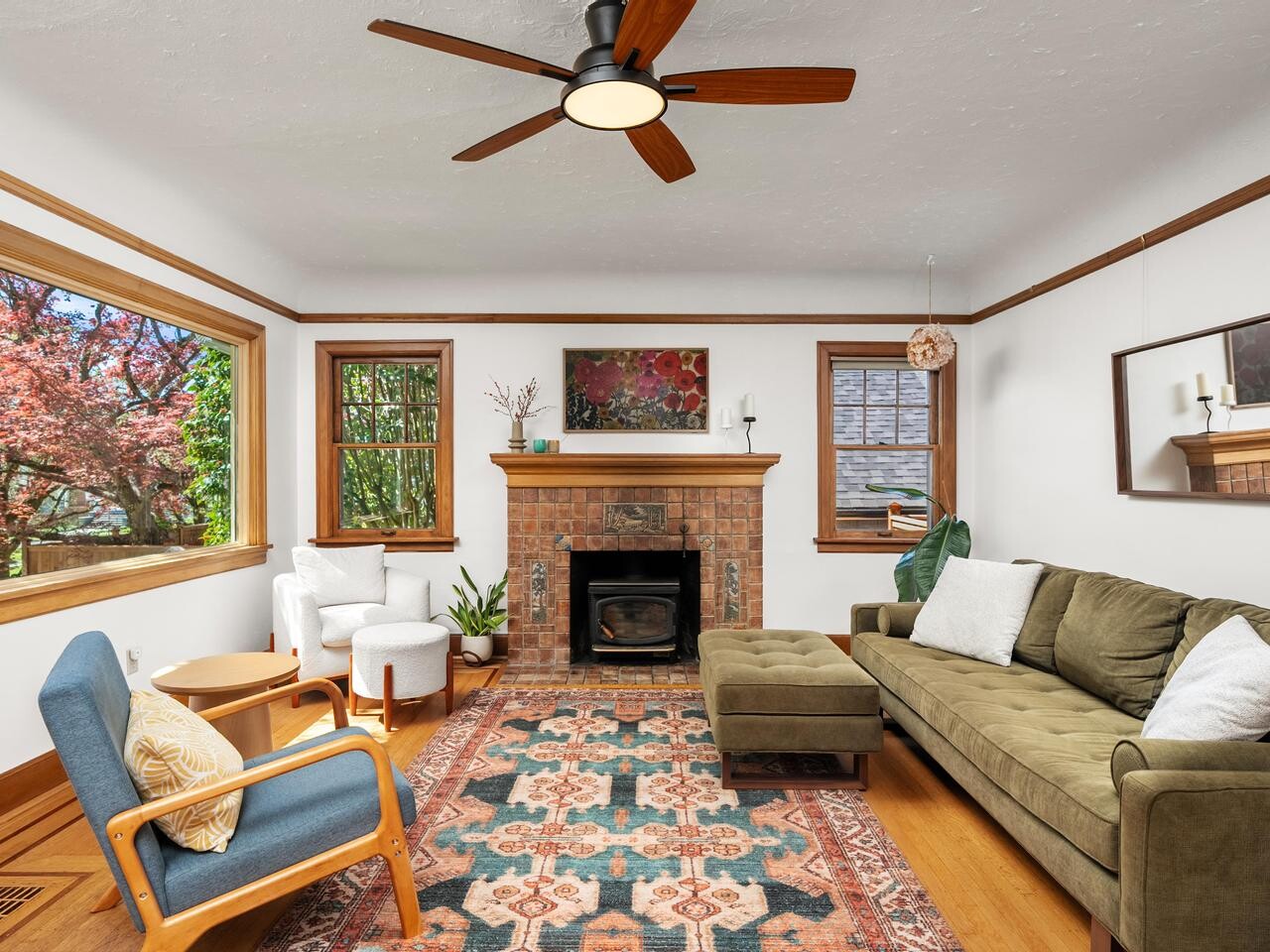 A cozy living room with a green sofa, armchair, large rug, fireplace, ceiling fan, and big windows showing trees outside. Plants and wall art add decorative touches to the bright, inviting space.