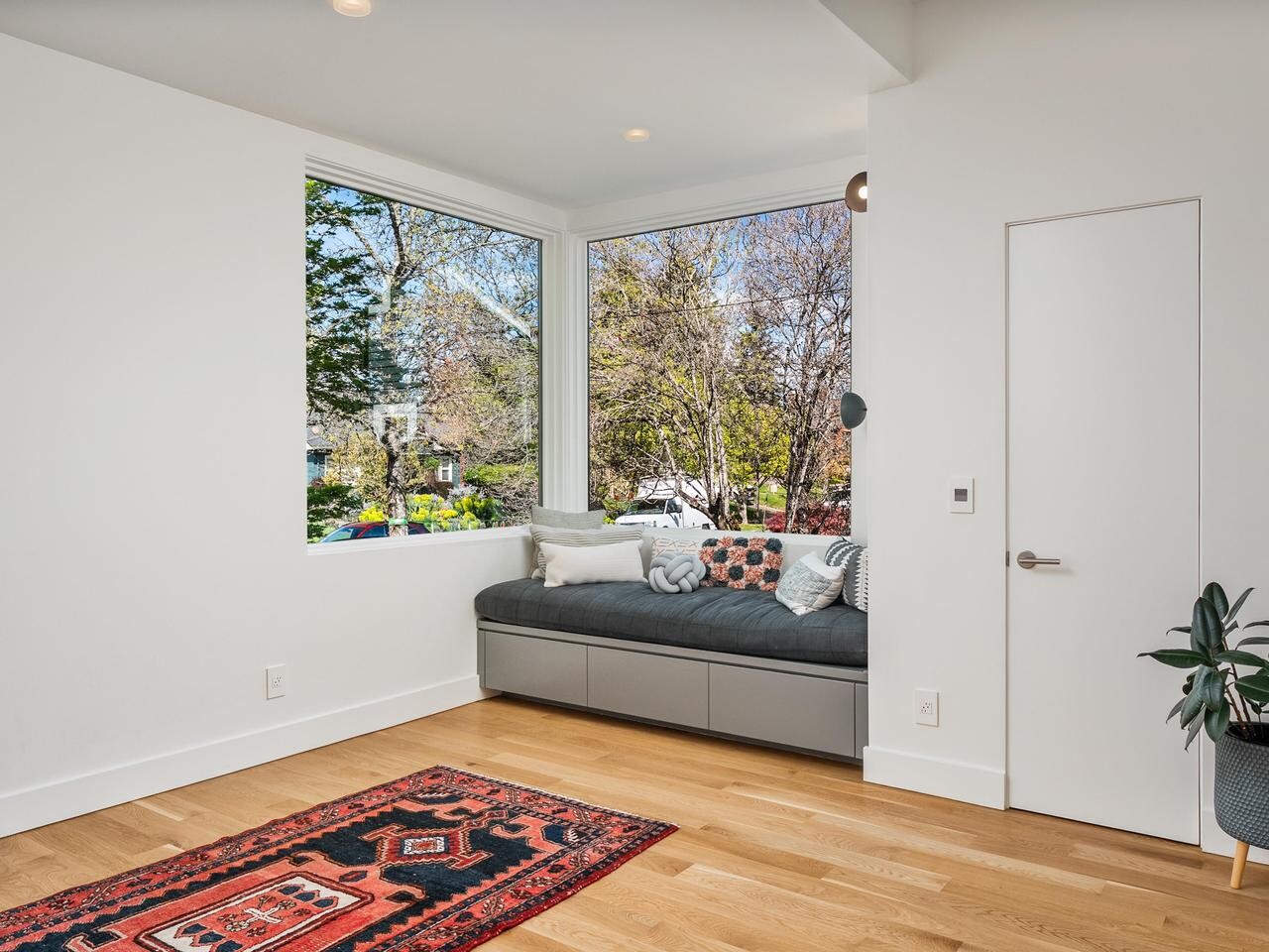 A cozy, modern room with a window seat filled with cushions, large corner windows showing trees outside, light wood floors, a red patterned rug, white walls, and a potted plant near a closed door.
