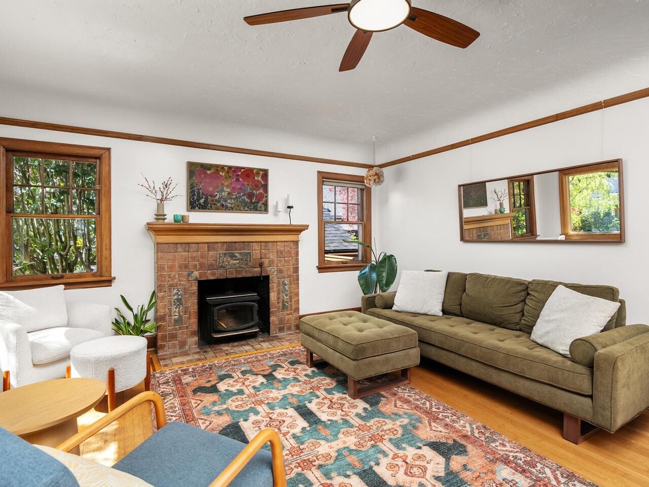 A cozy living room with a green sectional sofa, two white chairs, a blue accent chair, a brick fireplace, plants, and colorful art on the wall. Wood trim and large windows let in natural light.
