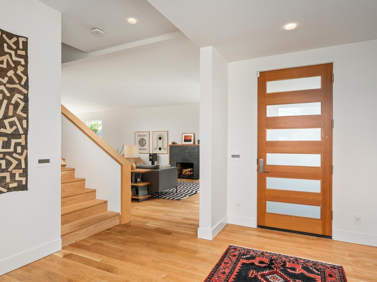 Modern entryway with a wooden front door featuring horizontal glass panels, light wood floors, a red patterned rug, open stairs, and a view into a living room with contemporary furniture and wall art.