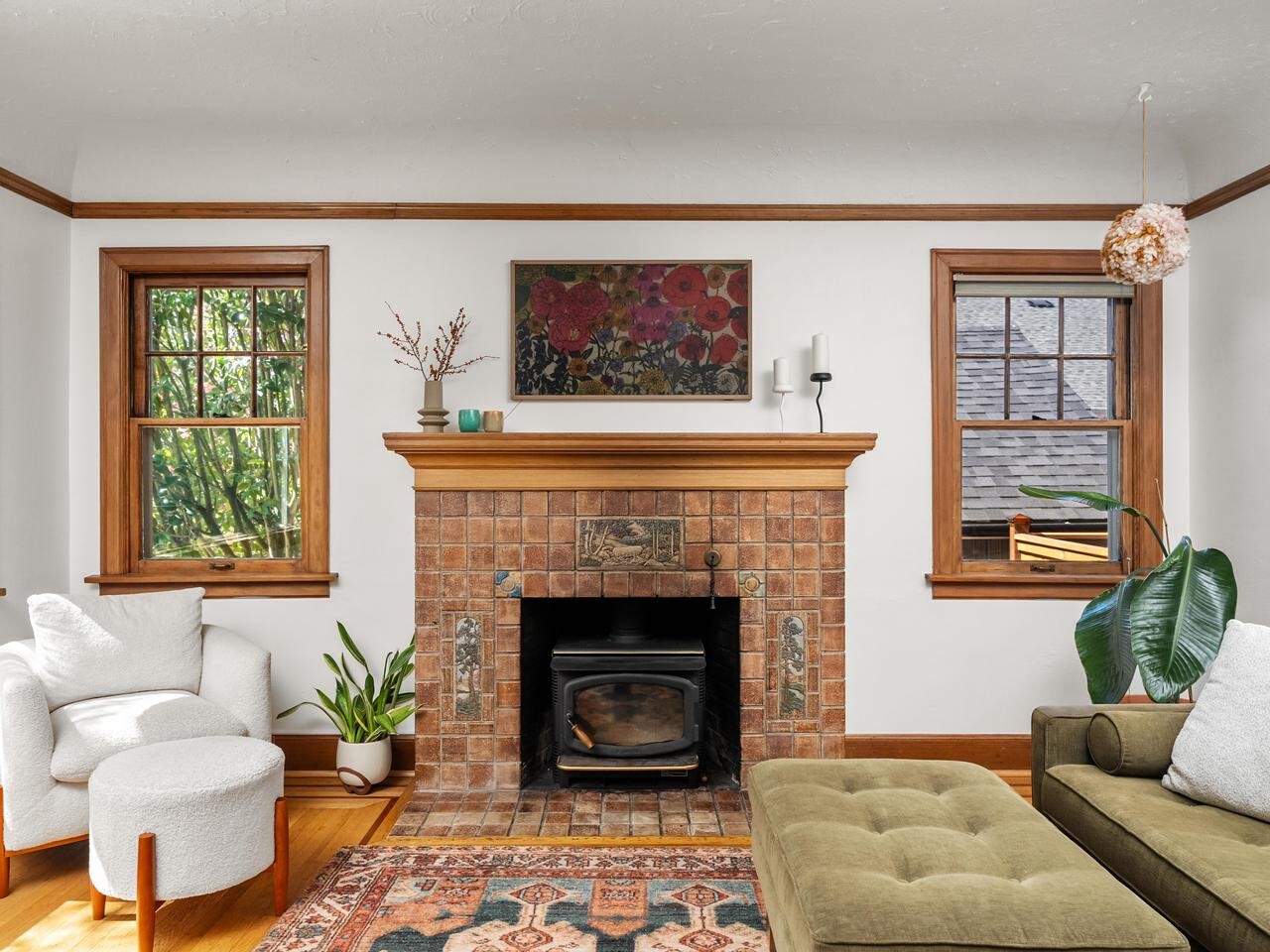A cozy living room with a brick fireplace, green sofa, white armchair, indoor plants, a patterned rug, and large windows allowing natural light to brighten the space.