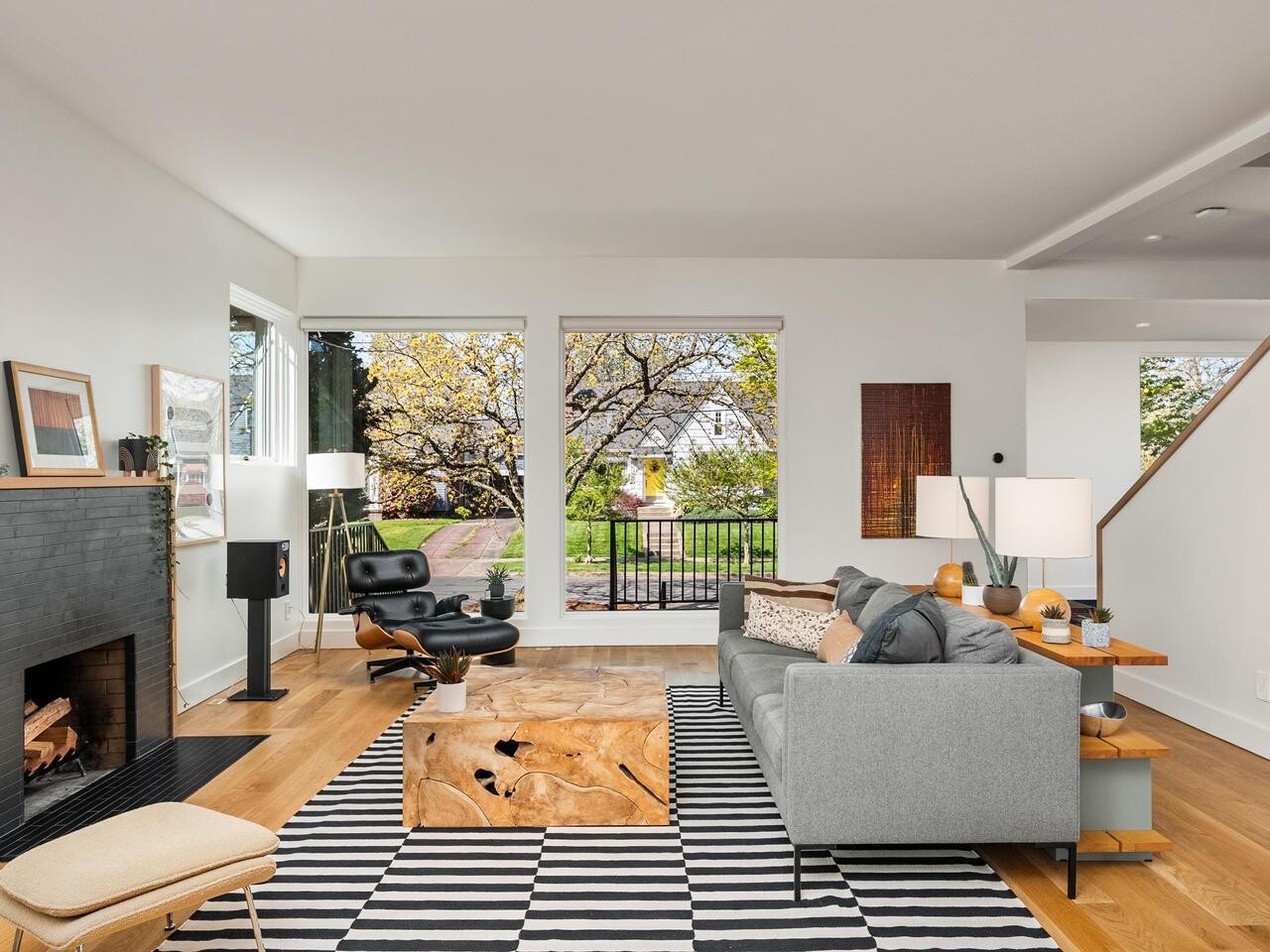 Modern living room with large windows, a gray sofa, black armchair, wood coffee table, striped rug, fireplace, and art on the walls. Outside, trees and greenery are visible through the windows.