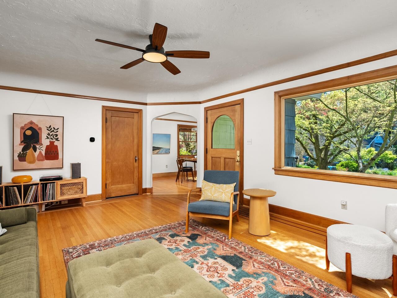 A bright, cozy living room with wood floors, a green sofa, colorful rug, mid-century furniture, large window with view of trees, and artwork on white walls. A doorway leads to a dining area.