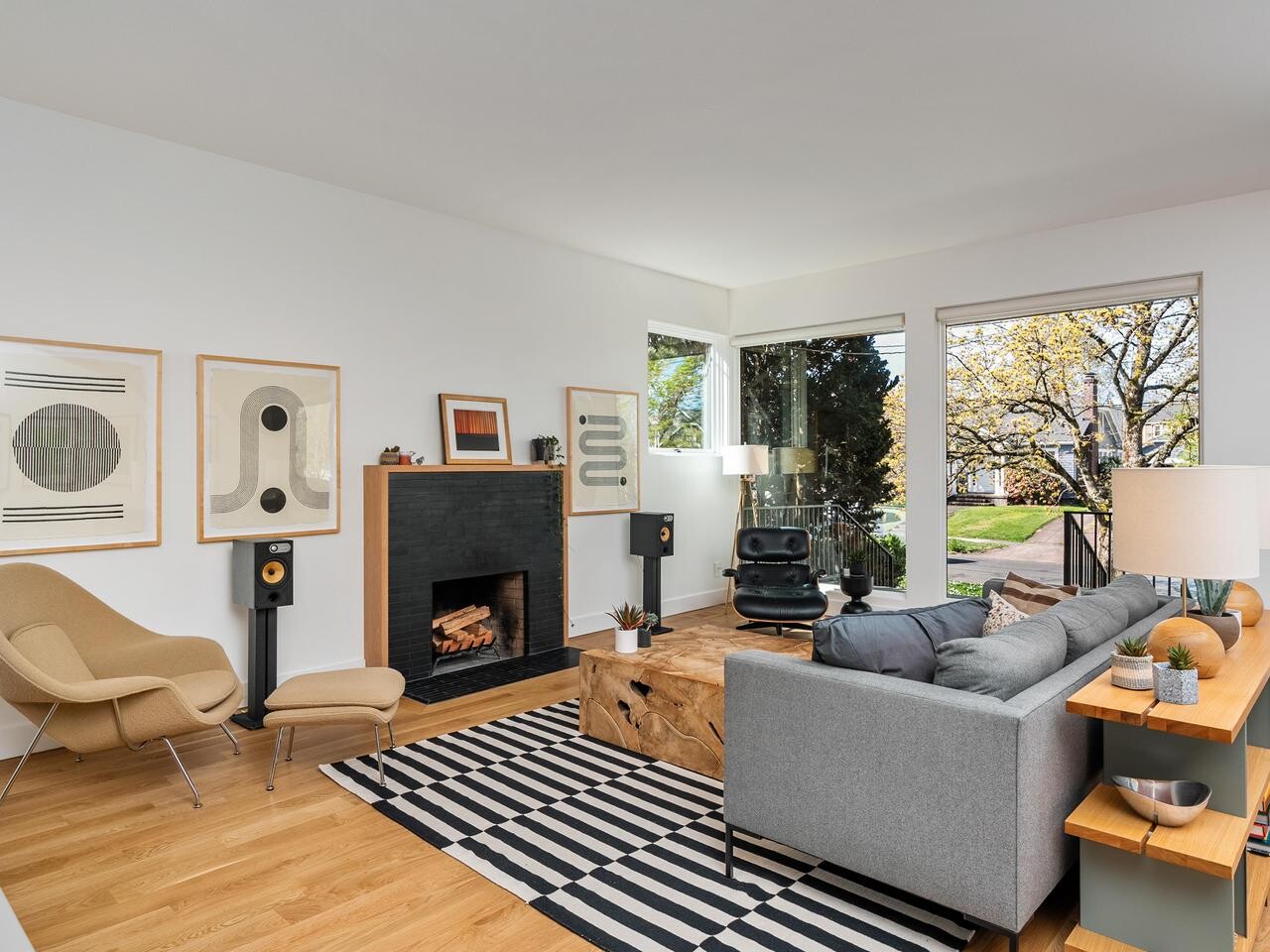 Modern living room with a gray sofa, wood coffee table, black accent chair, beige lounge chair, abstract art on white walls, striped rug, and large windows showing trees outside. Fireplace with stacked wood and decor on mantel.