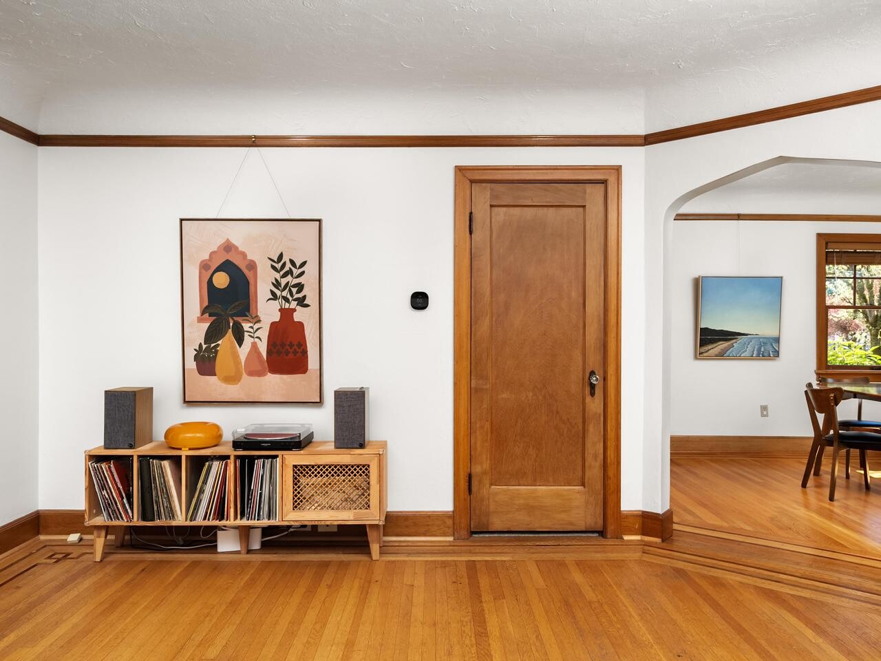 A cozy living room with wood floors and trim, a wooden door, and a low console holding records, speakers, and a turntable. A colorful abstract art piece hangs above. An adjacent dining room is visible through an arched doorway.