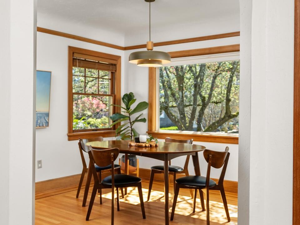 A bright dining room with wooden floors, a table set for four, modern chairs, large windows showing trees outside, a green plant in the corner, and a pendant light overhead.