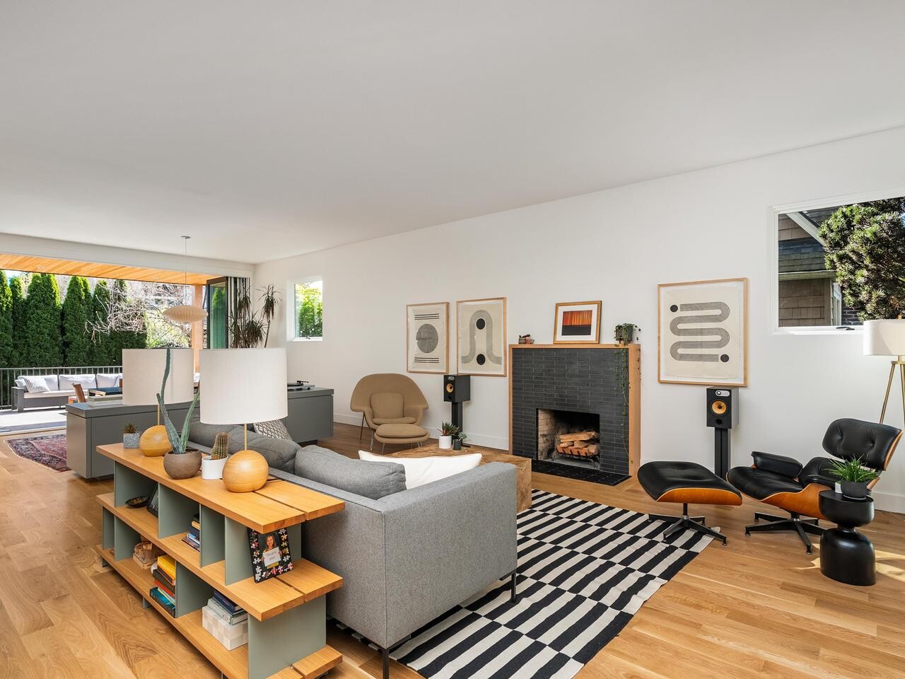 A modern living room with wood floors, a gray sofa, a striped rug, a black fireplace, lounge chairs, and large windows opening to an outdoor patio with greenery visible.