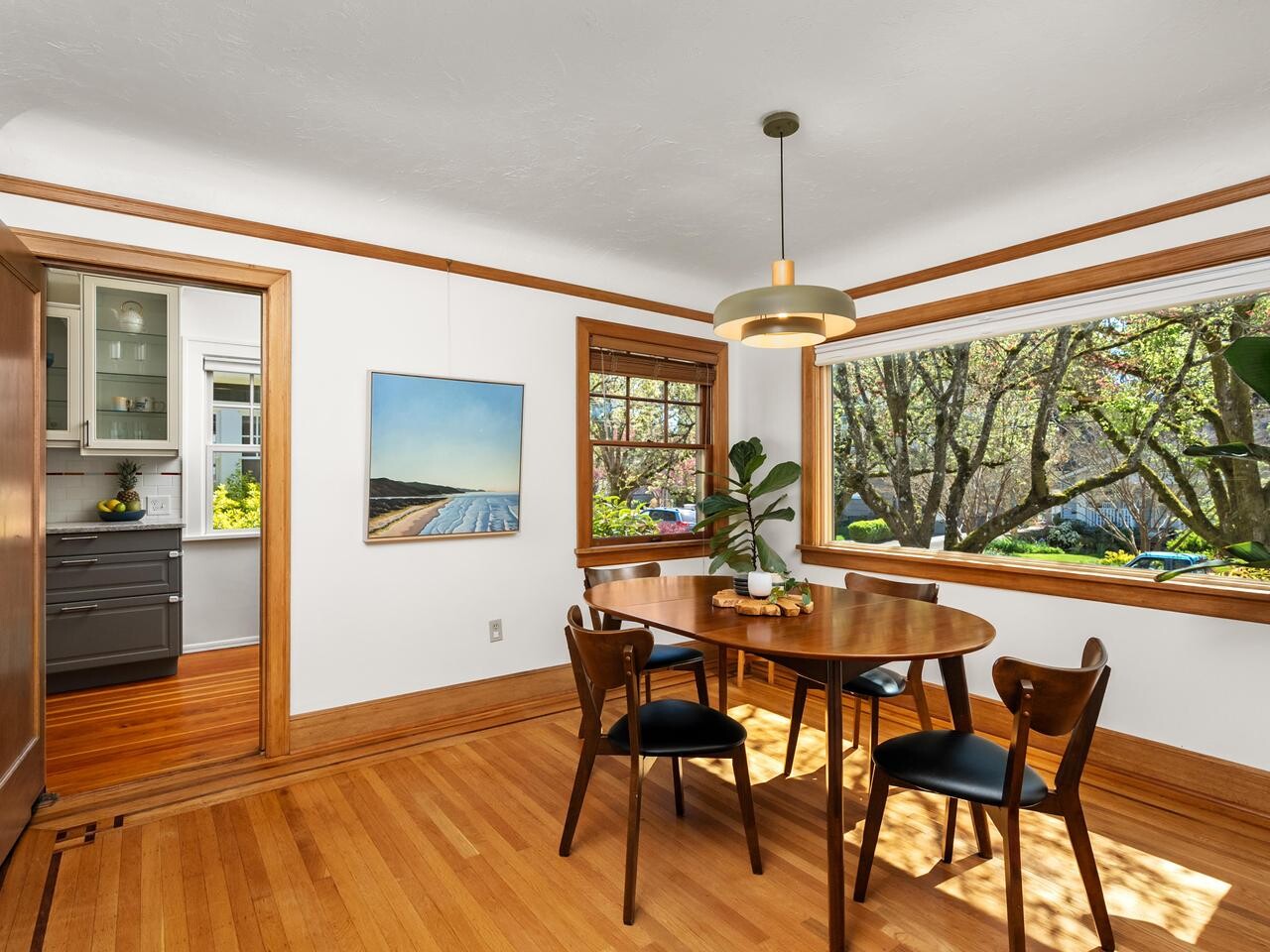 Bright dining room with a wooden table and four chairs, large window with a view of trees, wooden floors and trim, artwork on the wall, and a modern light fixture. Kitchen is visible through an open doorway.
