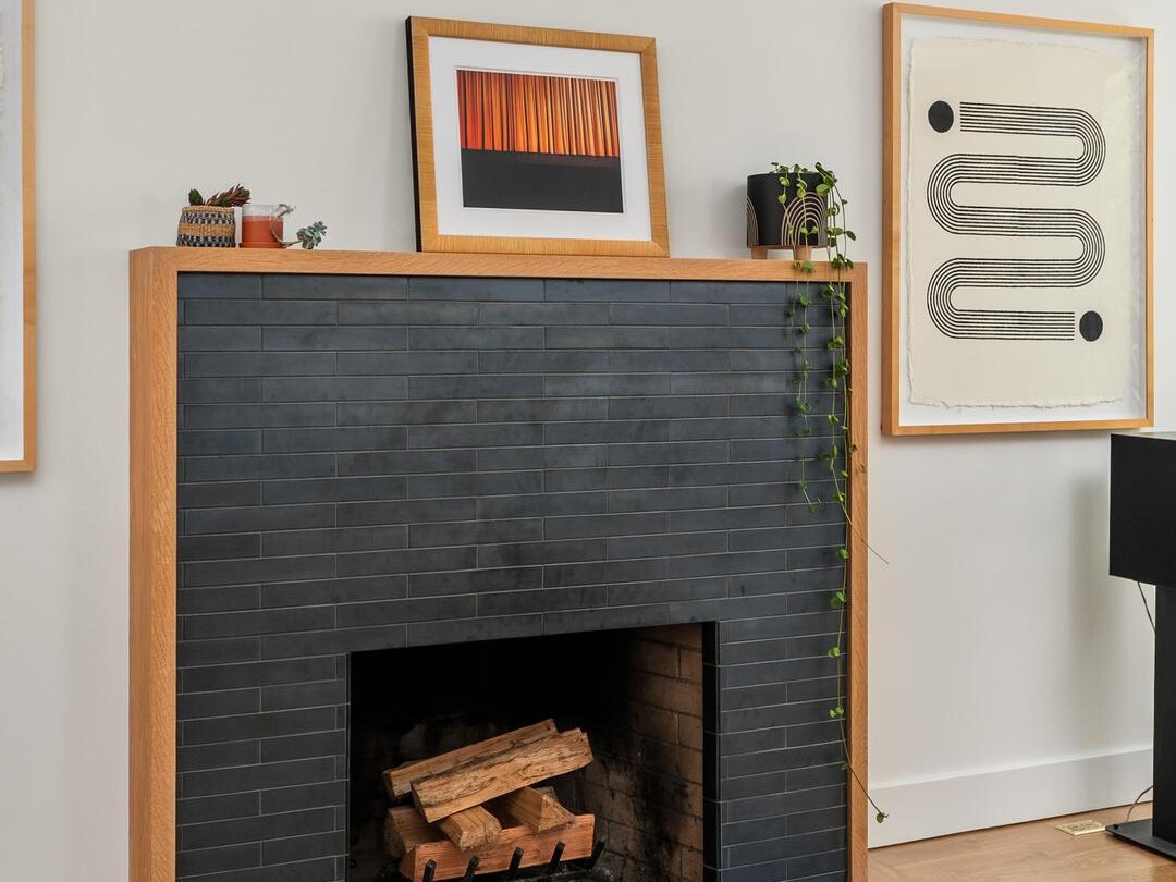 A modern fireplace with dark tiles and a light wood frame, topped with framed art and small potted plants. Geometric artwork hangs on the wall, and a wooden coffee table with succulents sits in the foreground.