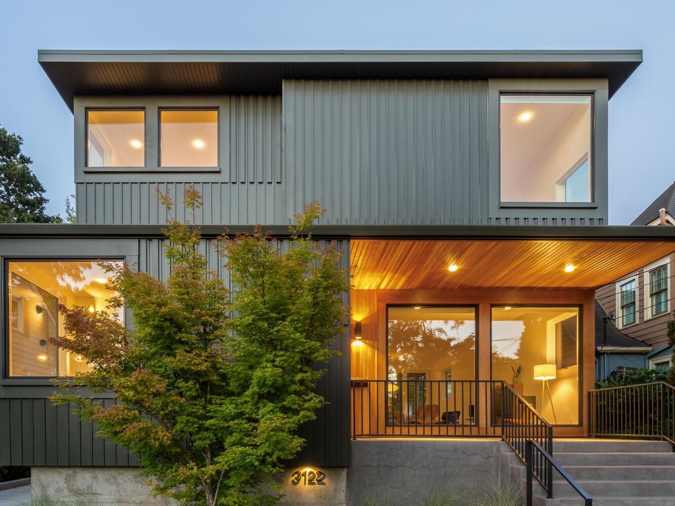 A modern two-story house with large windows, dark green vertical siding, and a wood-paneled porch ceiling. Warm indoor lighting is visible through the windows, and a small tree stands in front of the entrance steps.