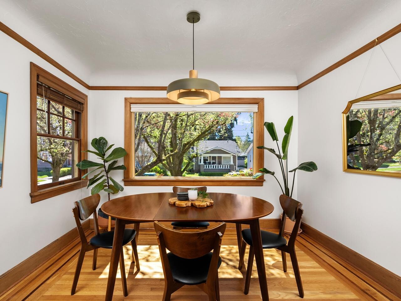 A bright dining room with a round wooden table and four chairs, large windows showing a green yard and house outside, indoor plants, a modern pendant light, wall art, and a gold-framed mirror.