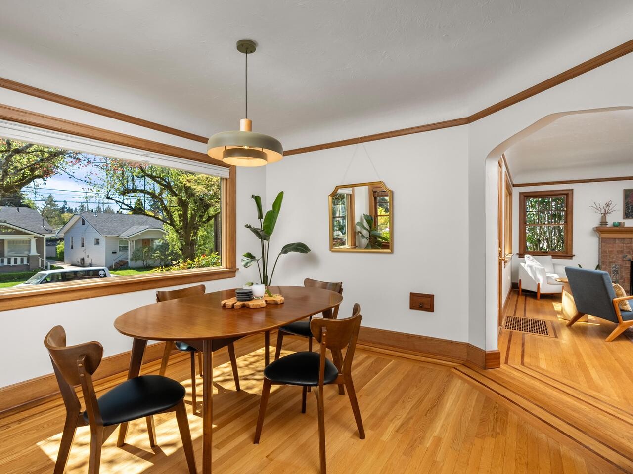 A bright dining room with a wooden table and four chairs, large window with a view of trees and houses, wooden floors, a pendant light, and plants. An archway leads to a cozy living room with a fireplace.